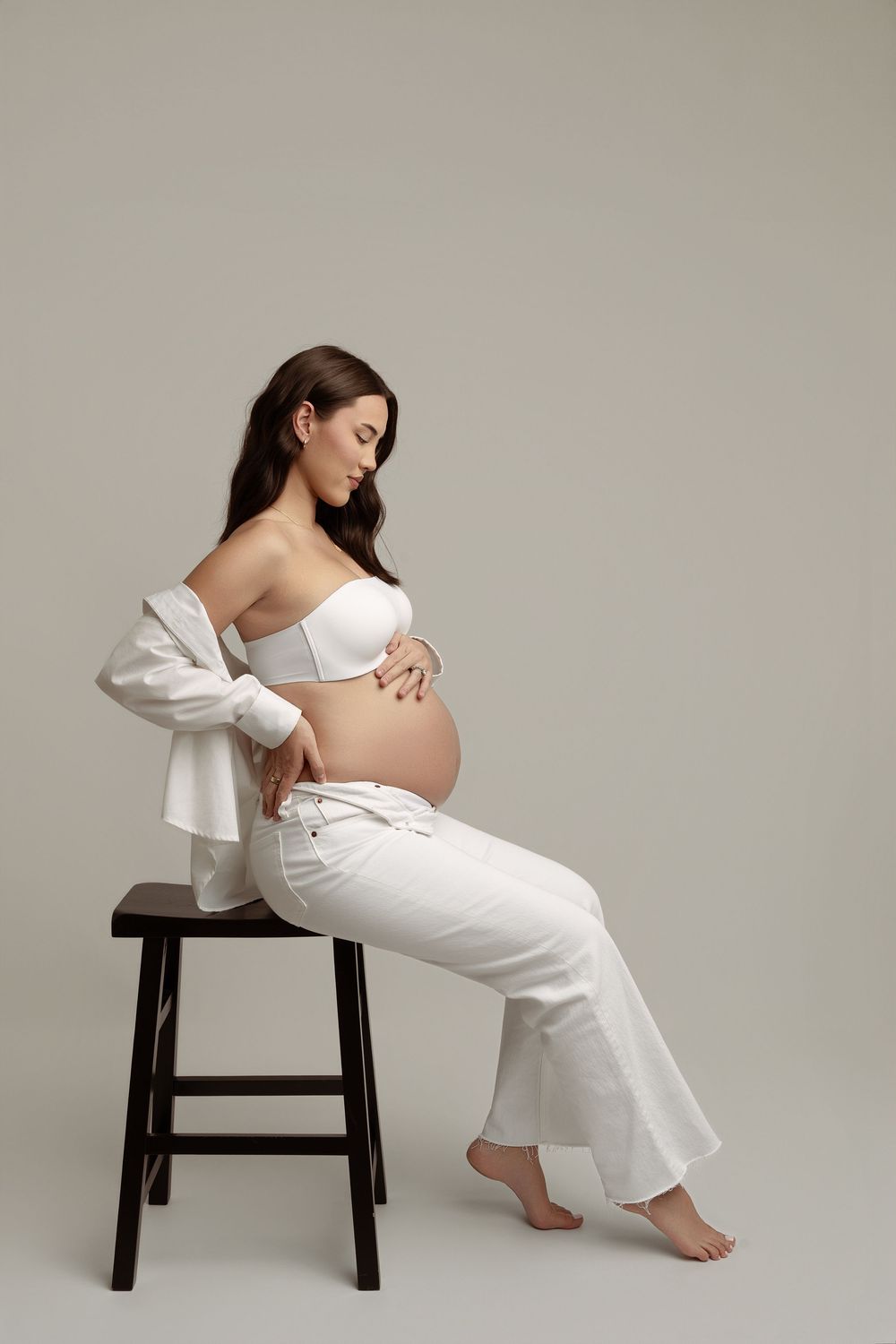 Pregnant woman in white strapless top and pants sits on stool, cradling baby bump against gray background.