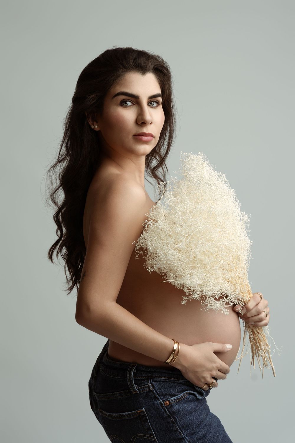 Pregnant woman in jeans holding a white dried floral bouquet, posing topless against a light gray background.