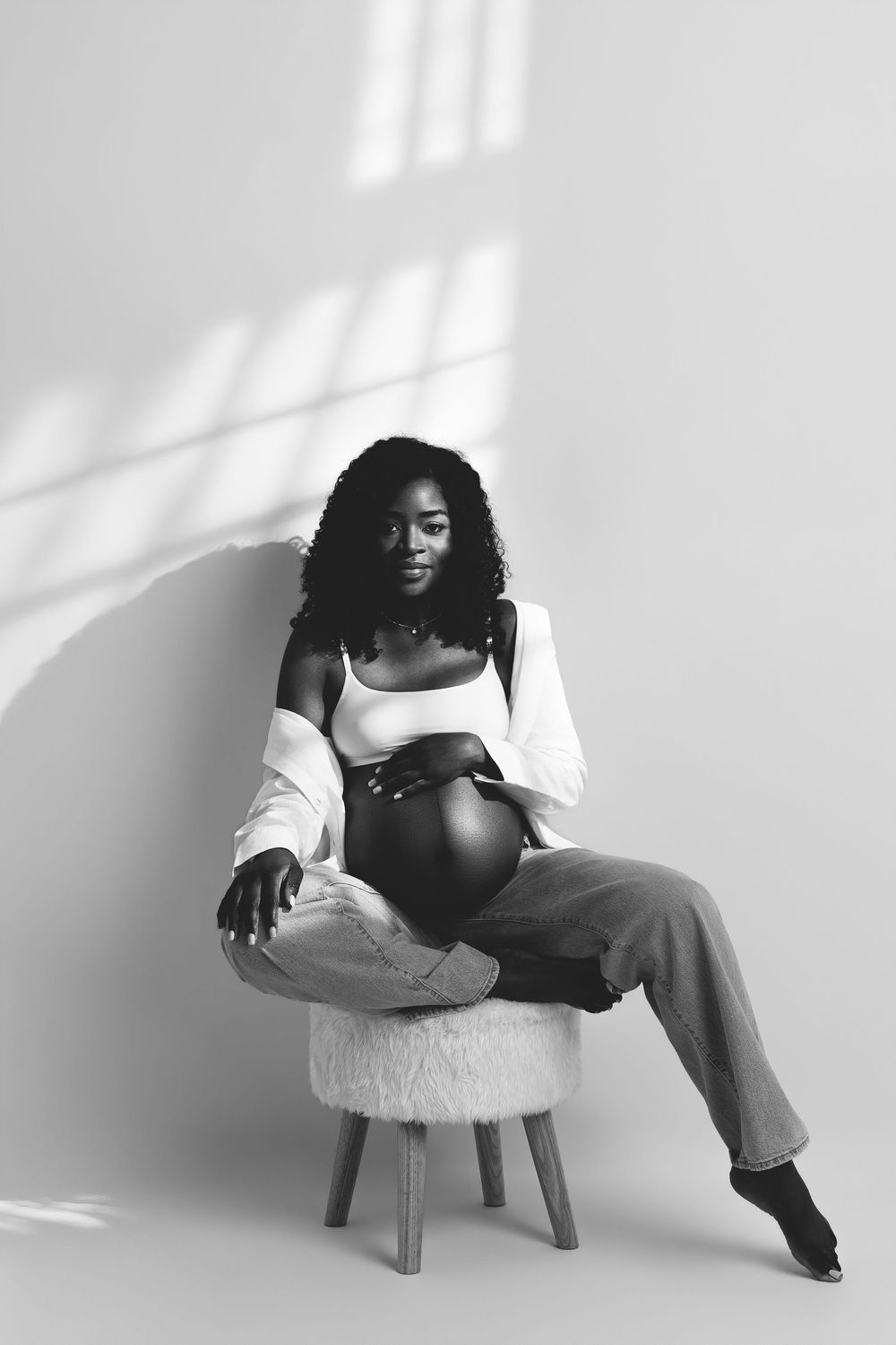 Black and white portrait of a pregnant Black woman sitting on a stool, cradling her baby bump in natural light.