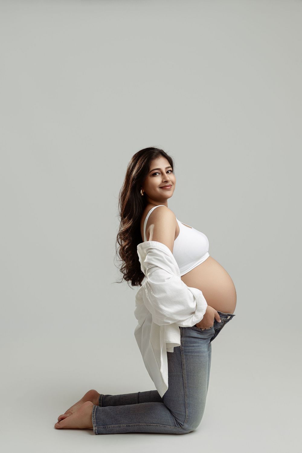 Smiling pregnant woman kneels in white crop top and jeans, showing baby bump in studio.