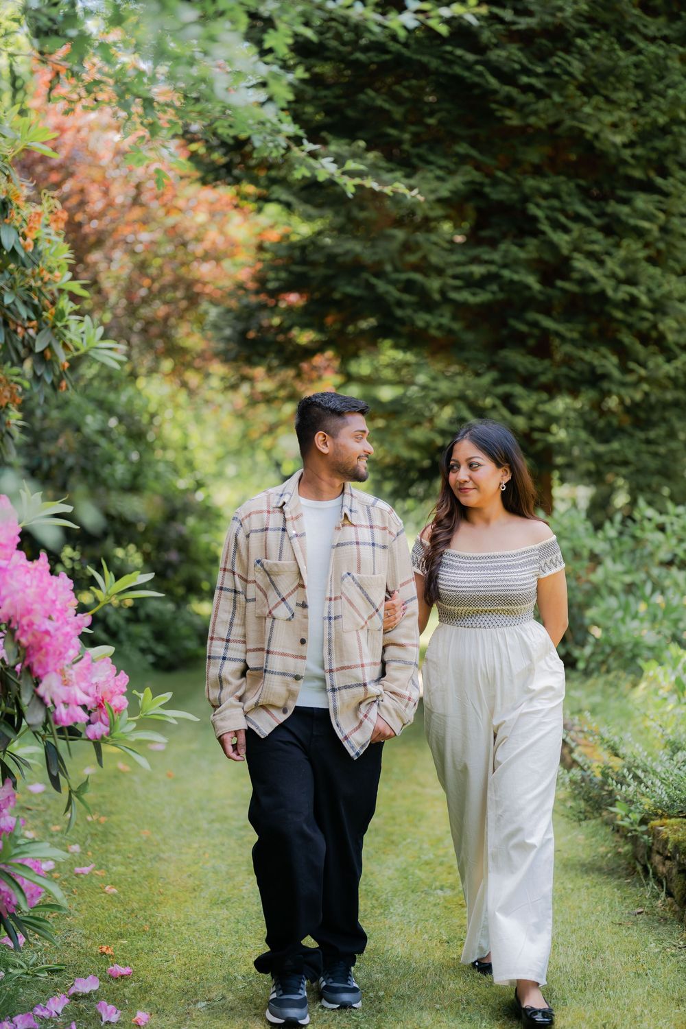 A couple walks hand in hand through a lush garden path, surrounded by pink flowers and green trees.