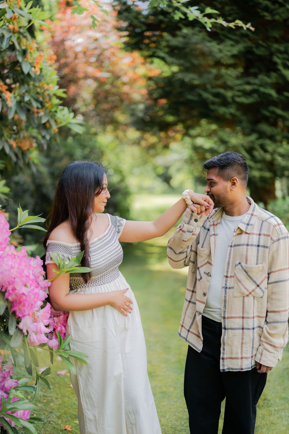 Pregnant woman in white dress holds man's hand in lush garden with pink flowers and green trees.