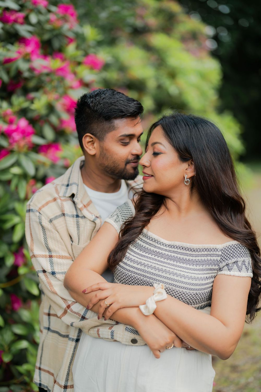 Romantic couple embracing near pink flowering bushes in a lush garden, woman in off-shoulder top, man in plaid shirt.