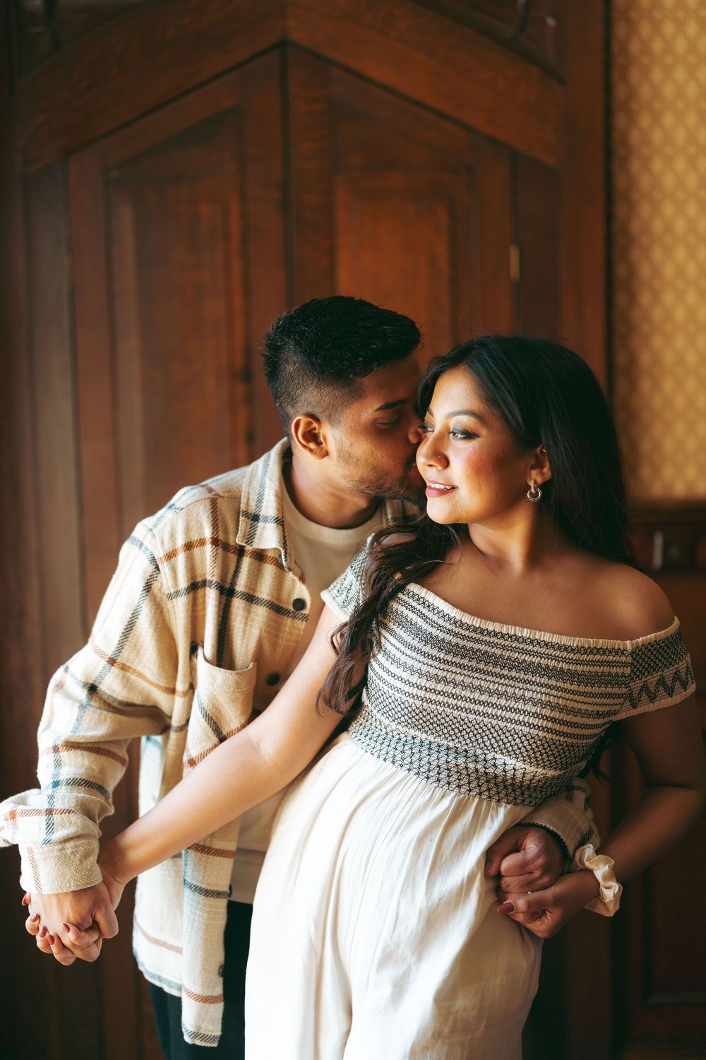 A couple shares a tender moment, man kissing woman's cheek while embracing indoors near wooden doors.