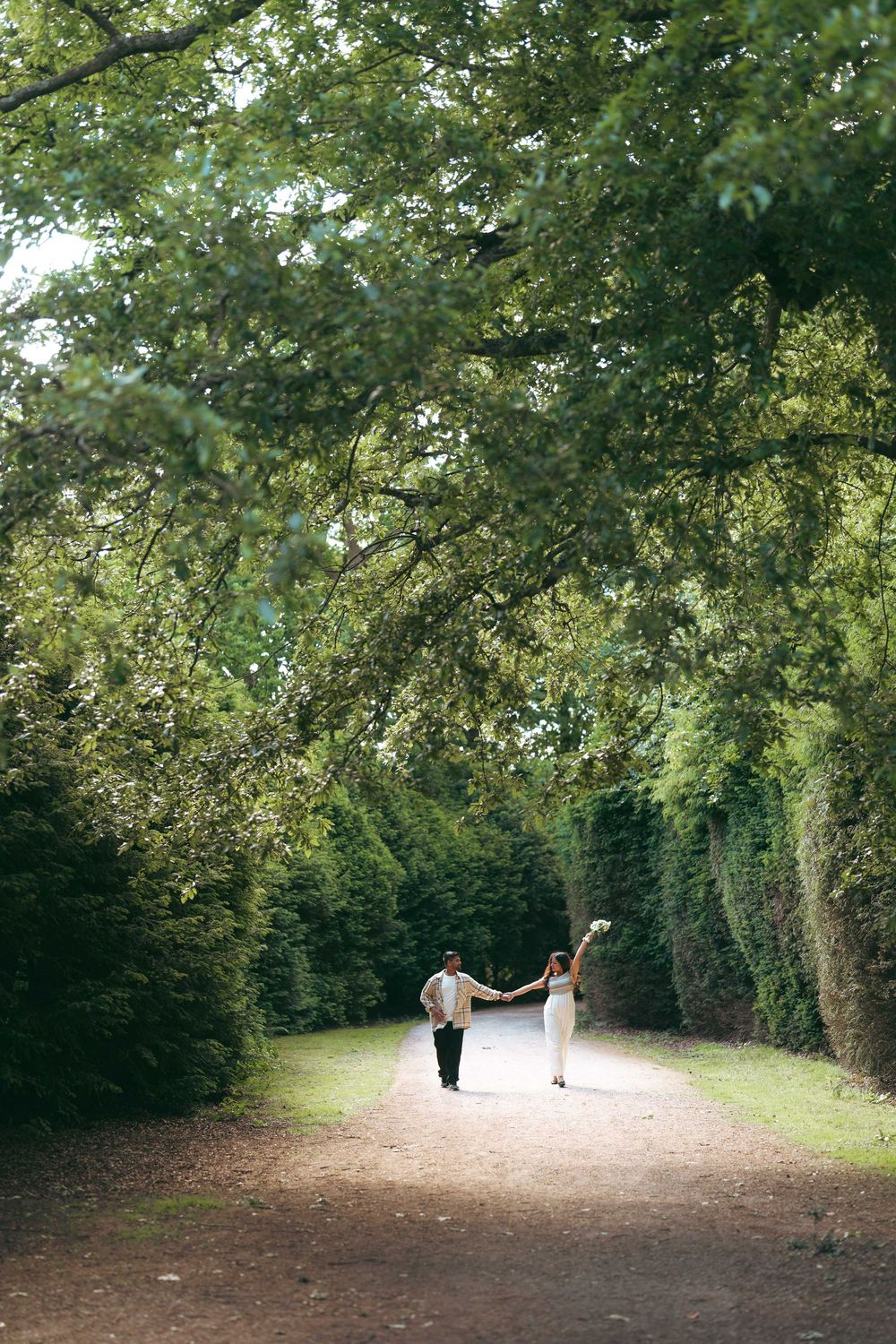 Couple holding hands walking down a tree-lined garden path surrounded by lush green hedges and foliage.
