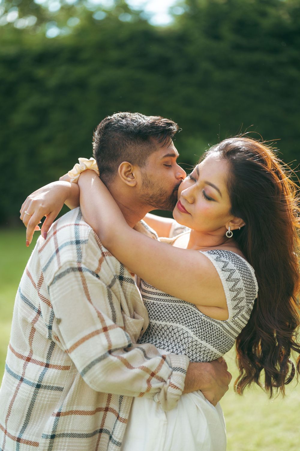 A couple shares a tender embrace and kiss outdoors, surrounded by lush green trees on a sunny day.