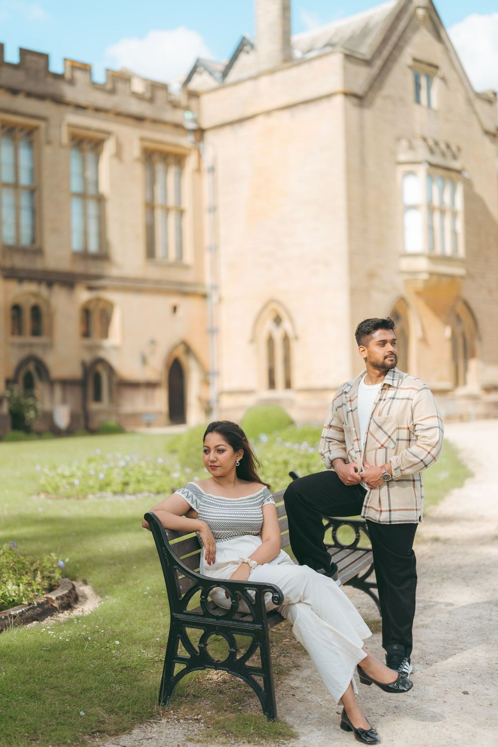 Couple posing at a historic stone castle, woman sitting on bench in white outfit, man standing in plaid shirt.