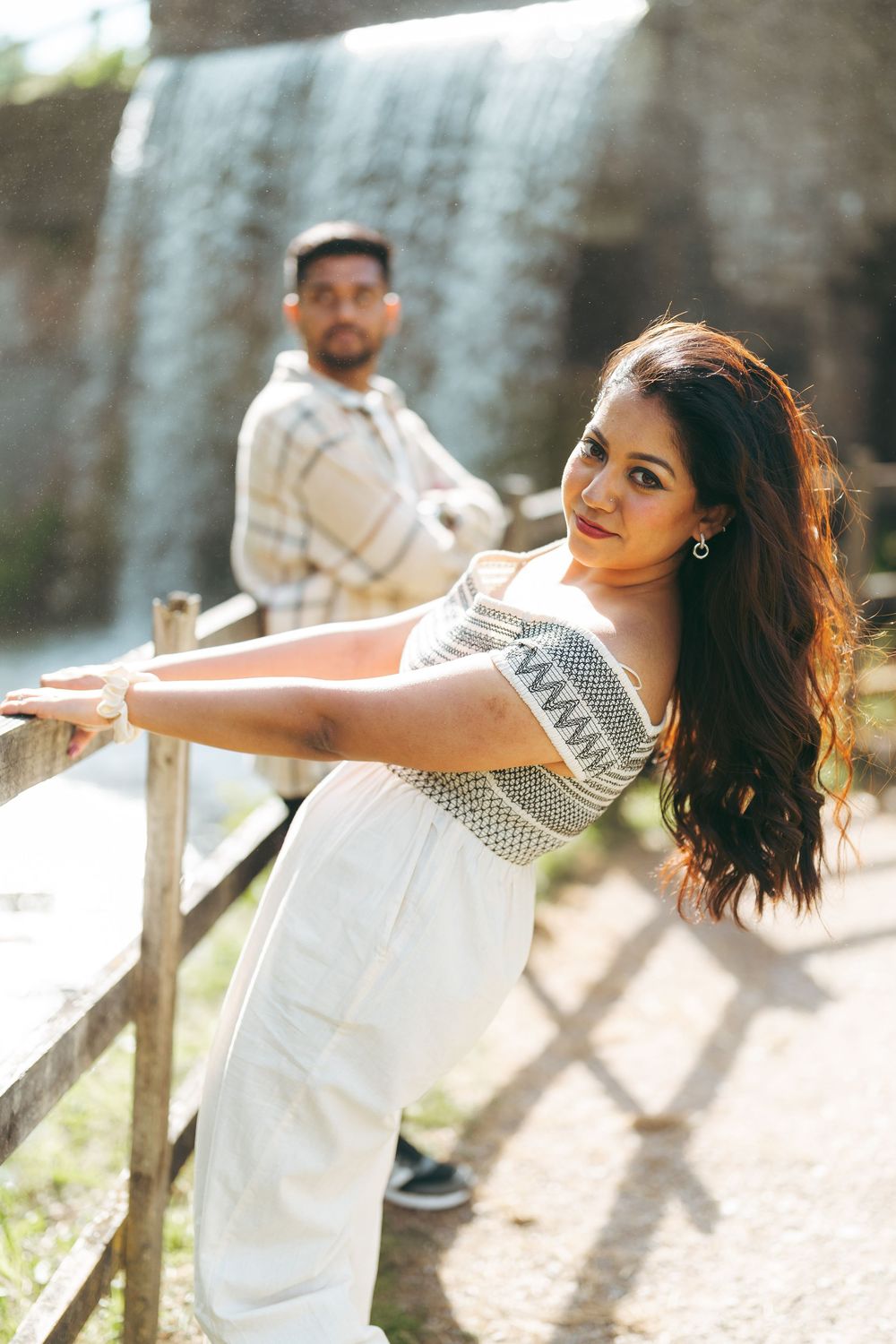 A woman in white dress poses at wooden fence with waterfall backdrop, man in plaid shirt stands behind her.