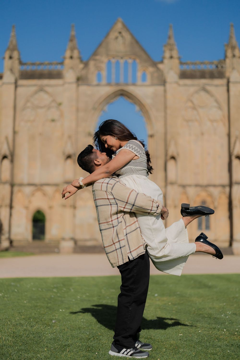 A couple shares a romantic lift and kiss in front of a historic Gothic stone cathedral on a sunny day.