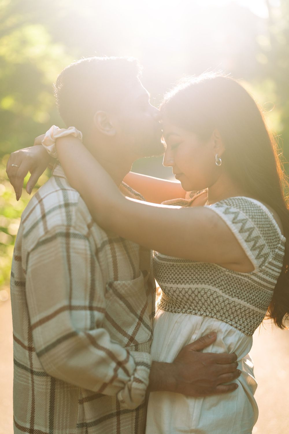Couple embracing romantically in golden sunlight outdoors, woman in patterned dress wrapping arms around man in plaid shirt.