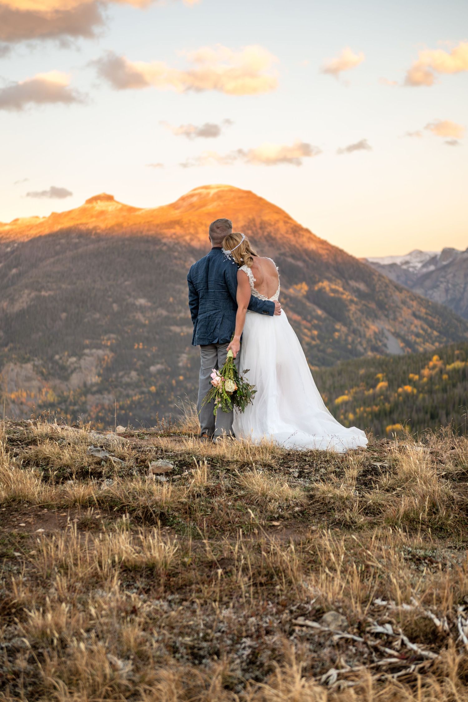 A bride holds her bouquet and lays her head on her grooms shoulder in the mountains with alpenglow on the mountain peaks surrounding them.