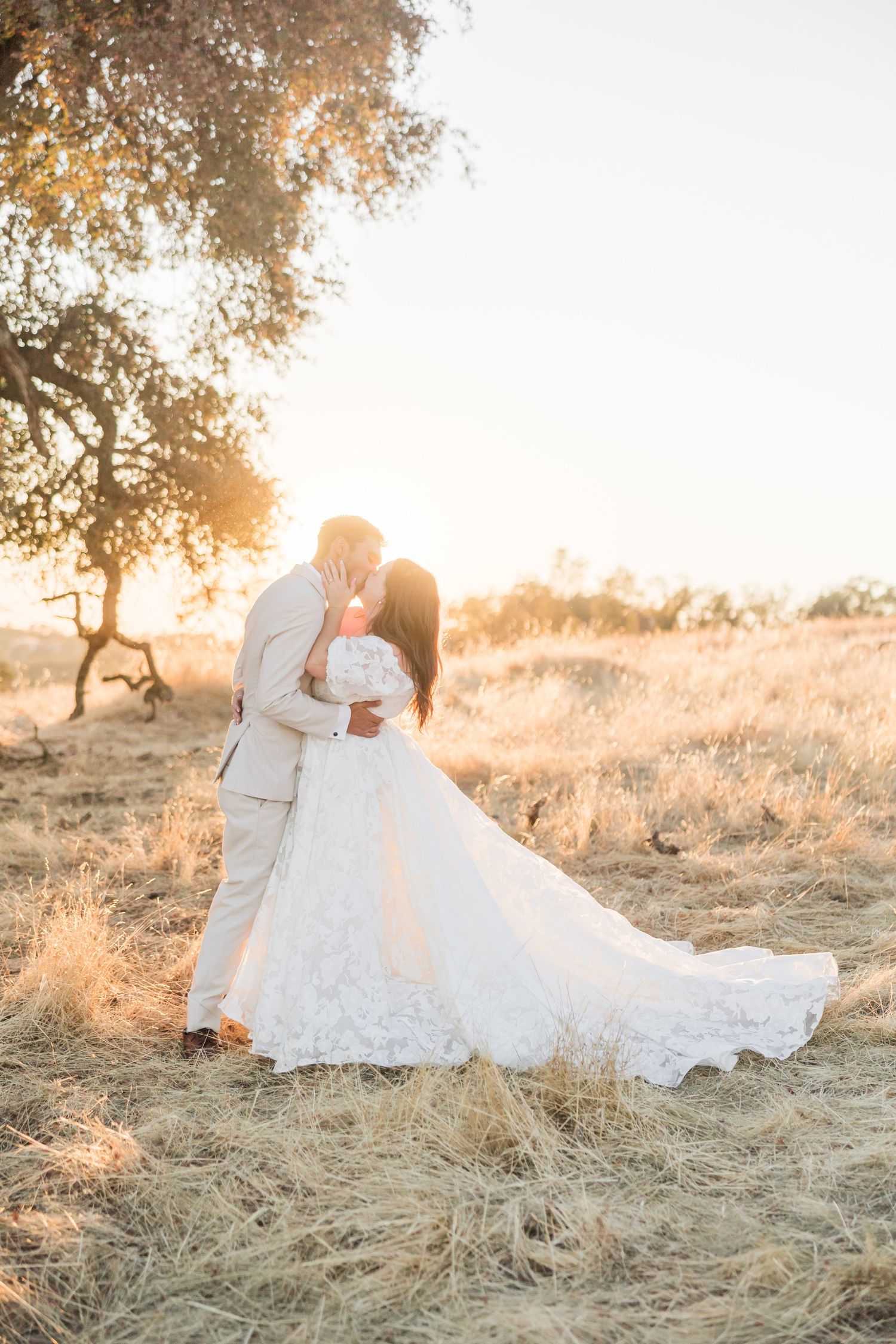 Bride and groom embrace at golden sunset under oak tree, bride in lace gown with long train holding bouquet.