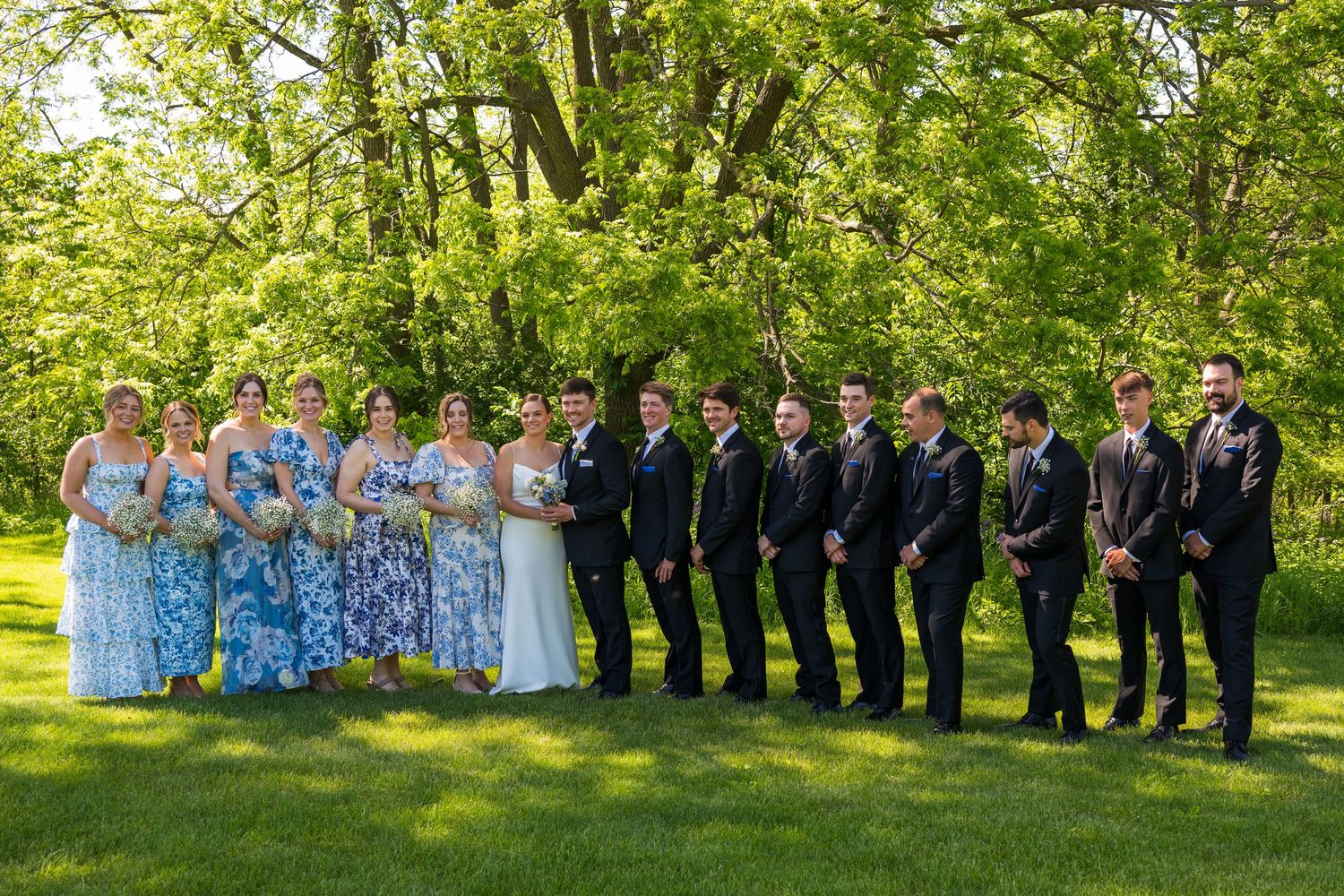 Wedding party poses outdoors on green lawn with bridesmaids in blue floral gowns and groomsmen in dark suits among lush trees.