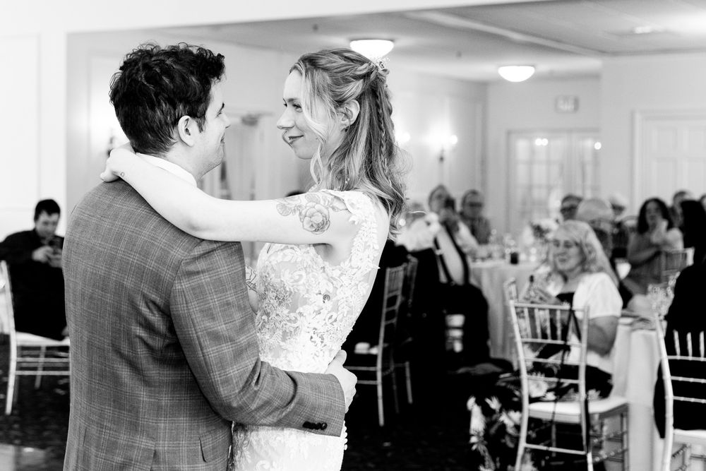 Bride and groom share their first dance, smiling joyfully, with wedding guests seated in the background.