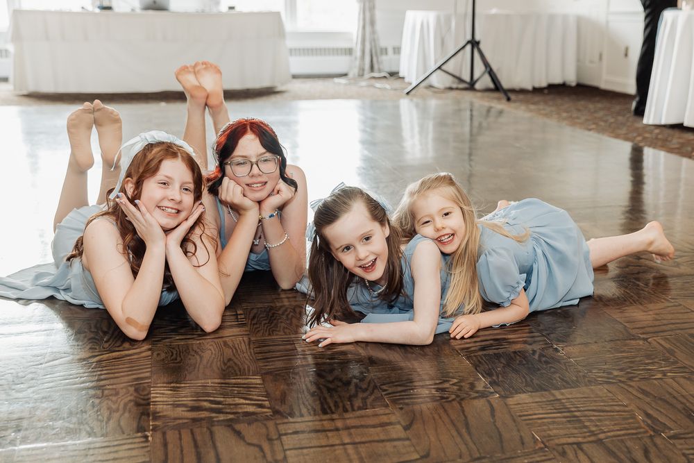 Four young girls in blue dresses lying on a wooden floor smiling, posing together for a dance recital photo.
