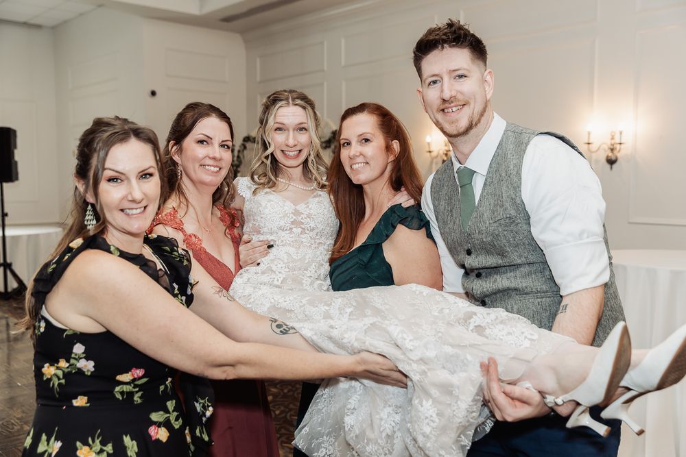Bride lifted by groom and bridesmaids smiling at wedding reception.