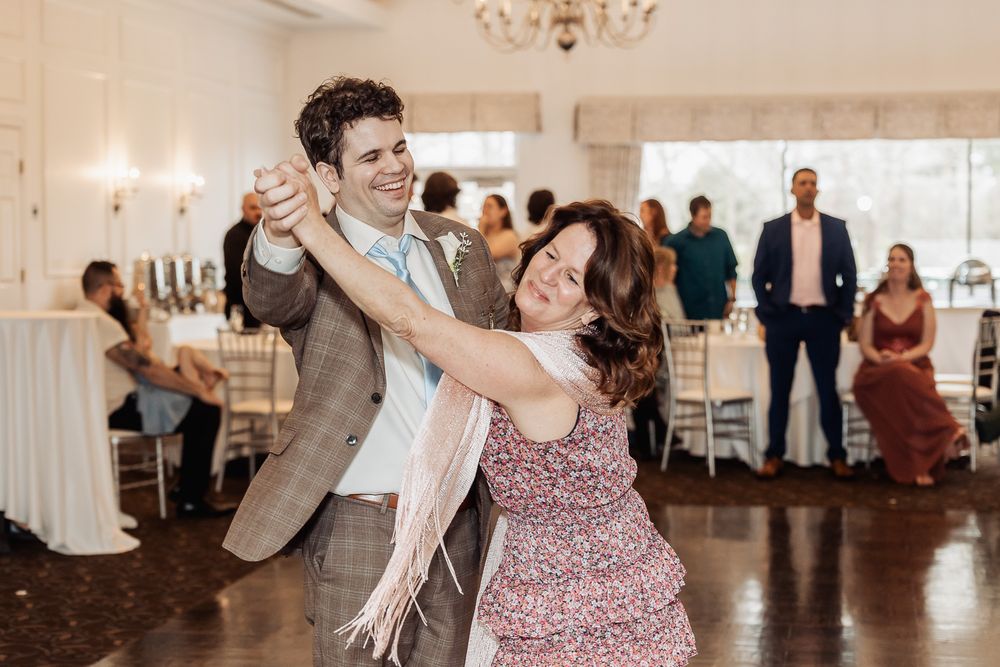 Groom dances with mother at wedding reception, smiling warmly, guests visible in elegant venue background.