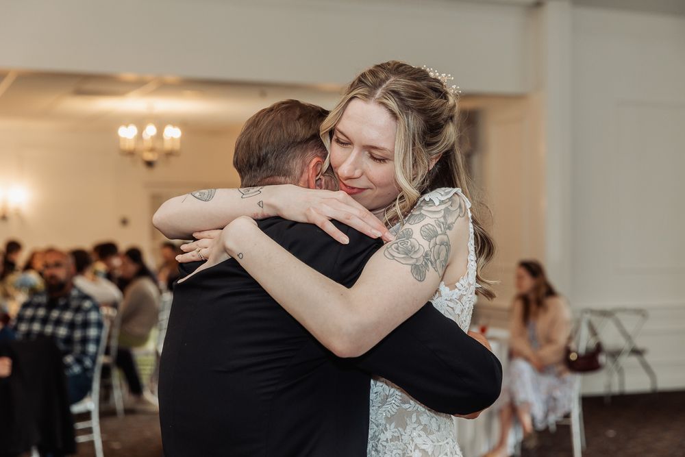 Bride in lace gown shares a heartfelt hug during a wedding reception dance in a bright ballroom.