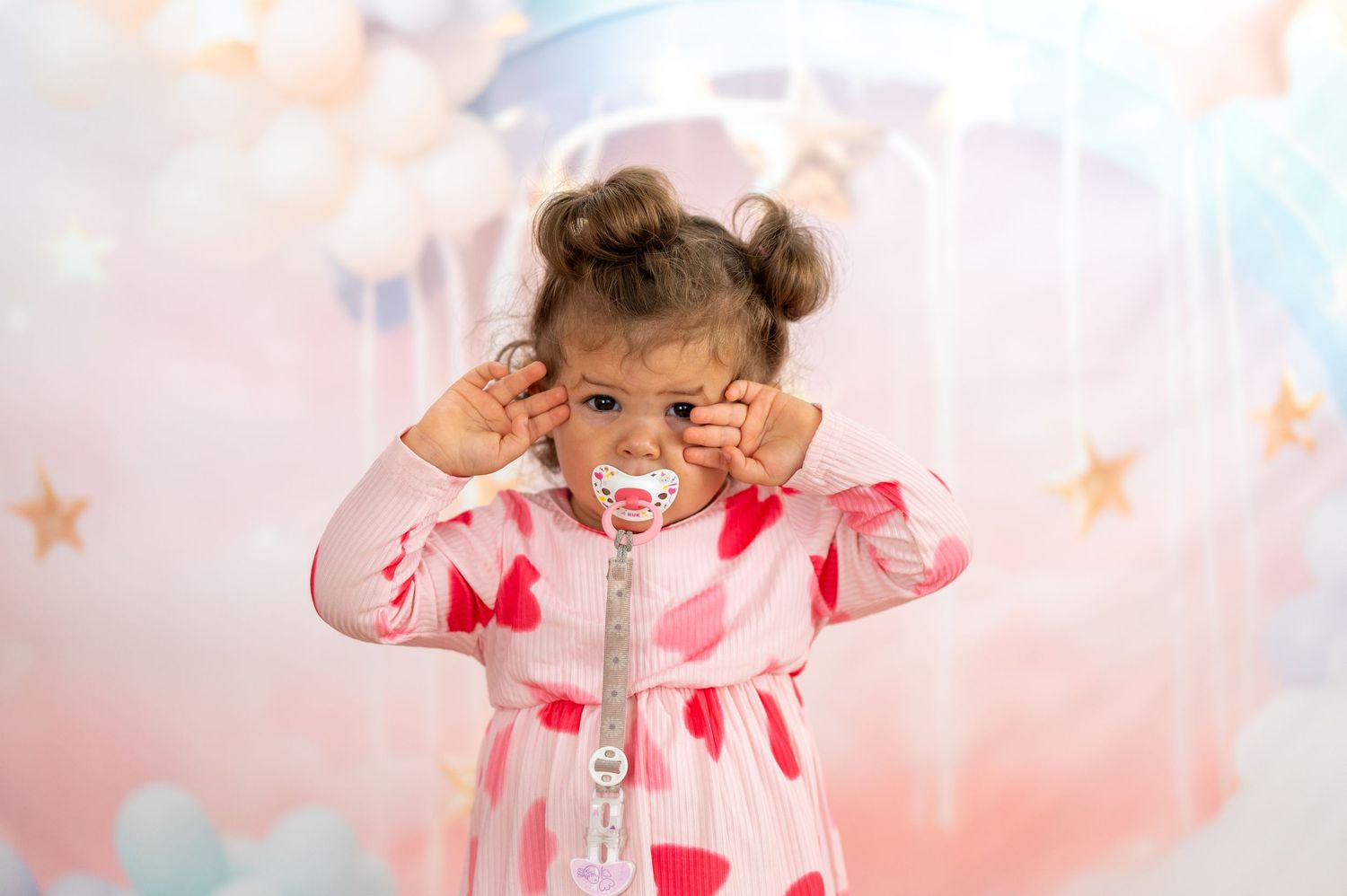 Tout-petit fille en robe de cœur rose avec des nattes et des poses de sucette devant une toile de fond de ballon pastel.