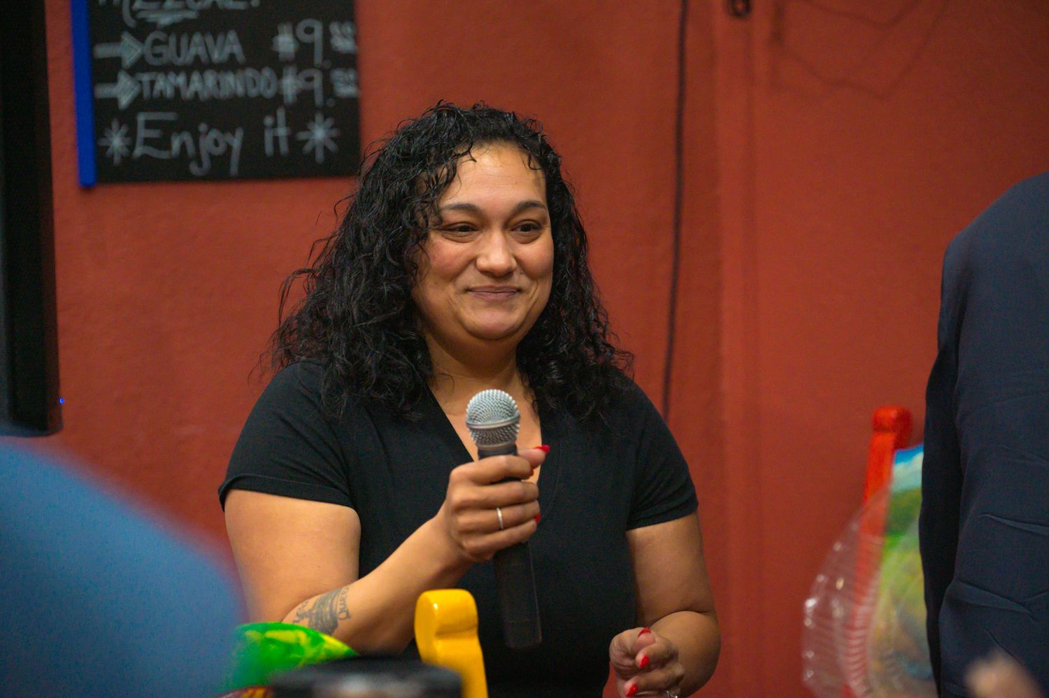 Woman with curly hair smiling and holding a microphone at an indoor event.