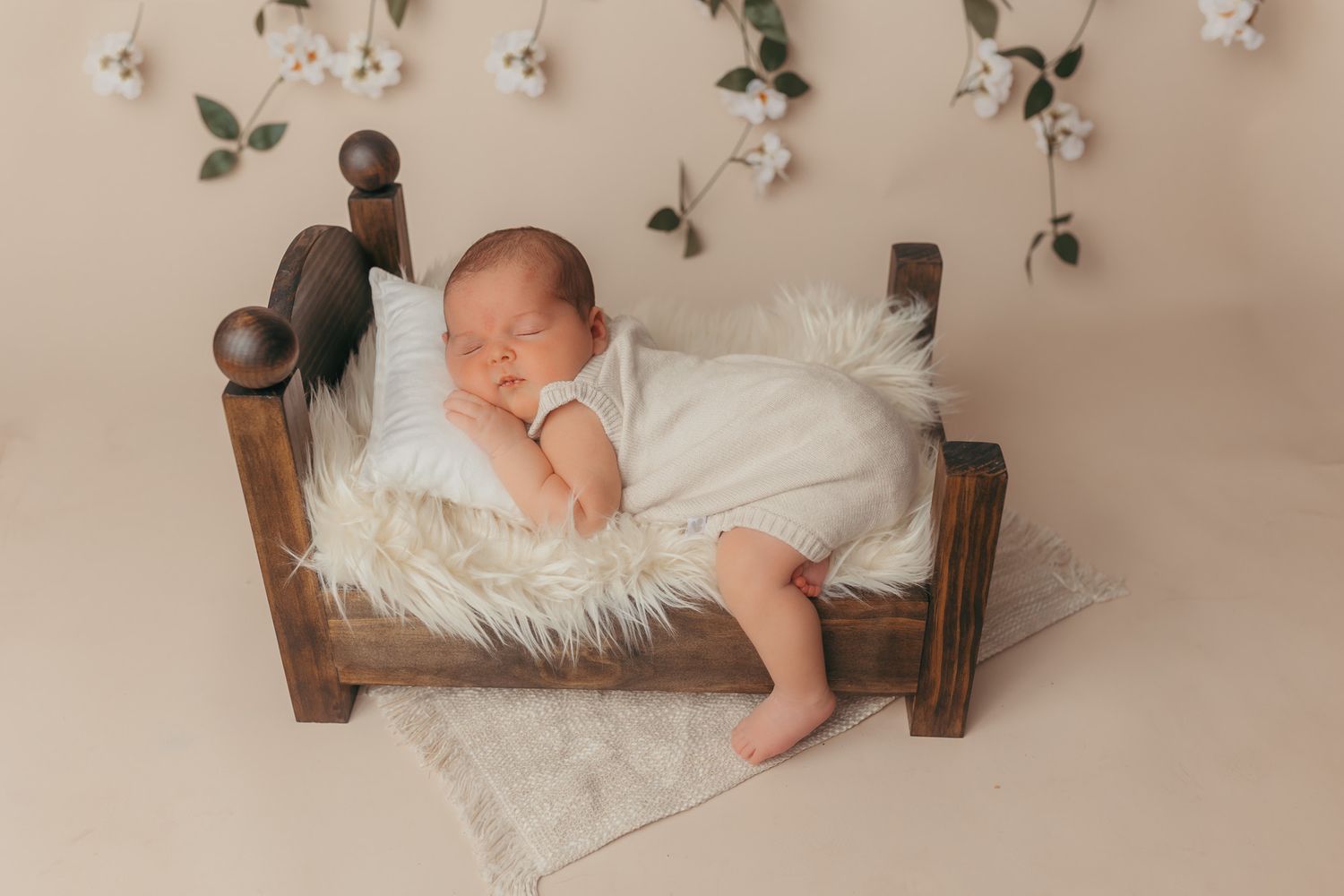 Newborn baby sleeping peacefully on a fluffy white fur blanket in a tiny wooden bed with floral backdrop.