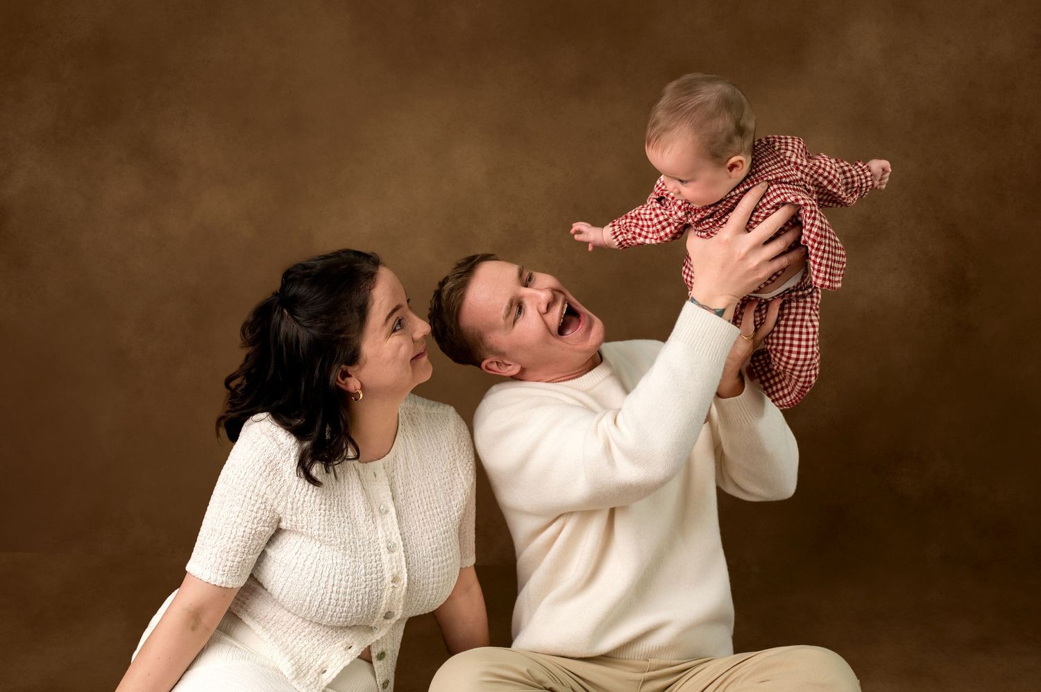 Happy family portrait with parents in white sweaters lifting smiling baby in red outfit against brown background.