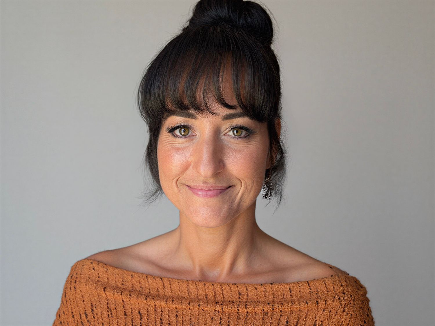 Smiling woman with dark hair in a bun, bangs, and green eyes wearing an off-shoulder rust orange knit sweater.