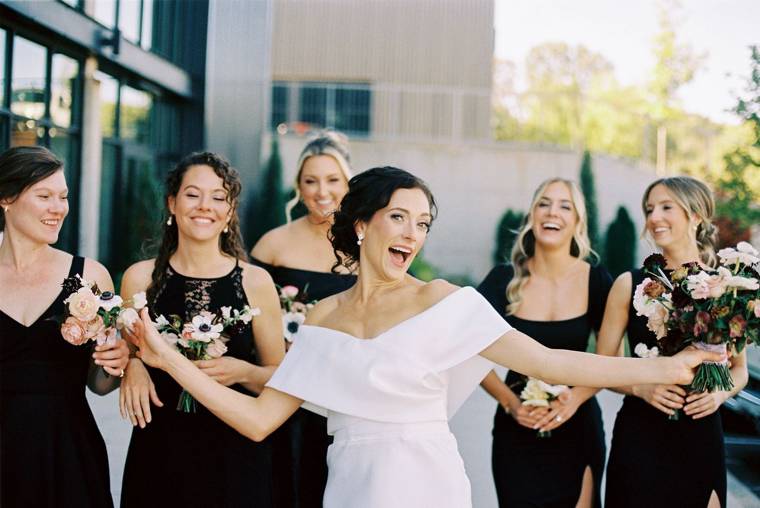 Bride in white gown laughing with bridesmaids in black dresses holding floral bouquets at outdoor wedding celebration.