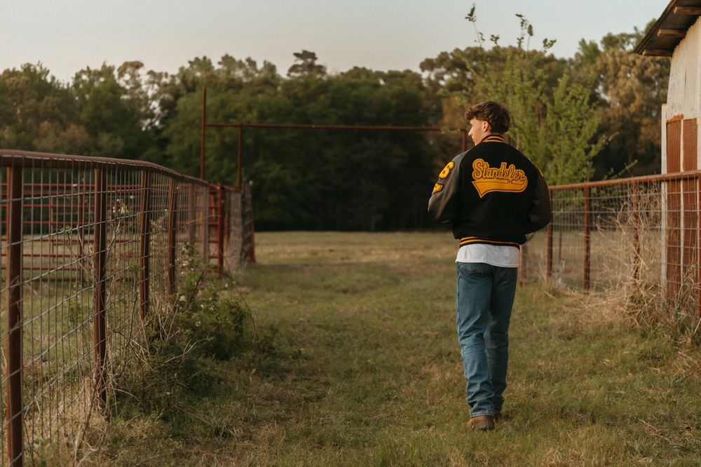 Teen in black jacket walking between rustic wire fences in grassy farm field at dusk.