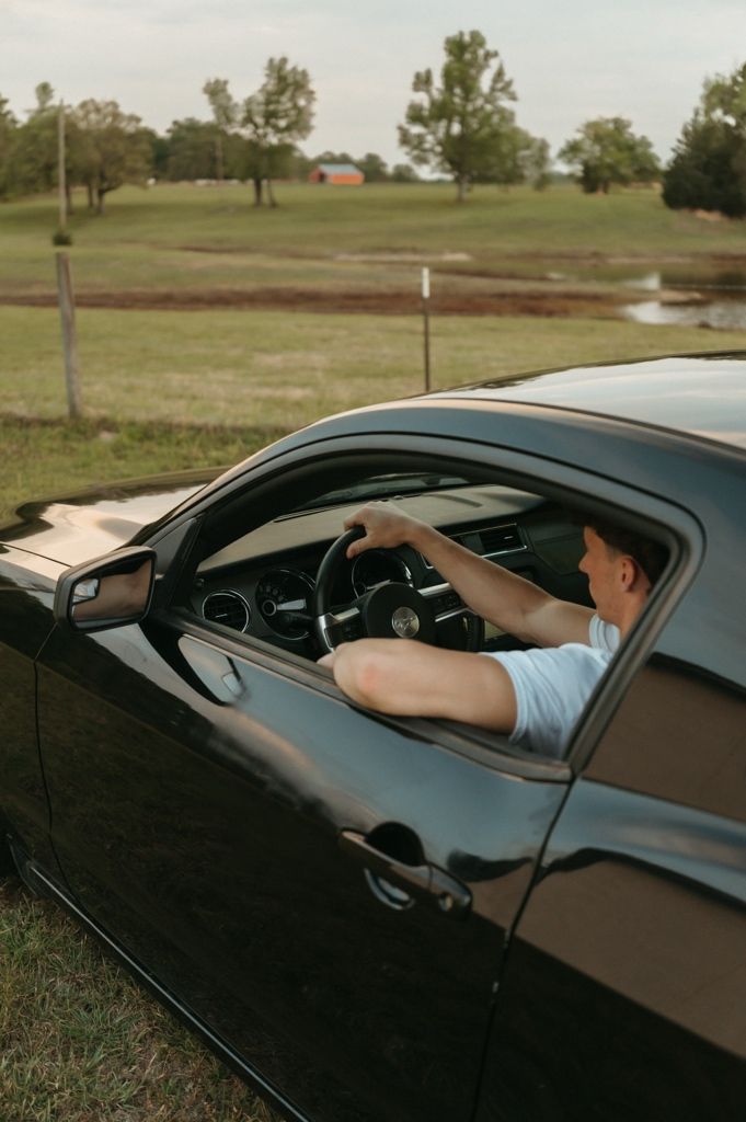 Young man in white shirt driving a black Ford Mustang on a rural farm road.