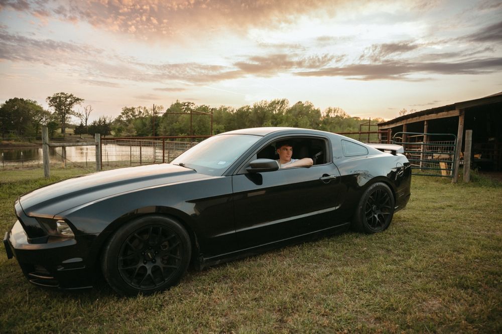 Teen boy sitting in a black Ford Mustang on a farm at sunset.