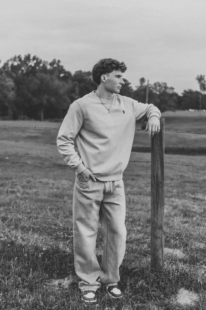 Young man in sweatshirt and jeans leans on a fence post in a rural field in black and white photo.