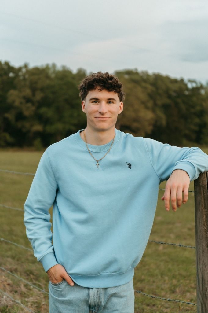 Young man in light blue sweatshirt leaning on fence post in rural field with trees.