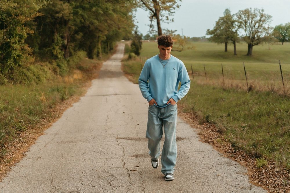 Young man in light blue sweatshirt and baggy jeans walking down a rural country road surrounded by green fields.