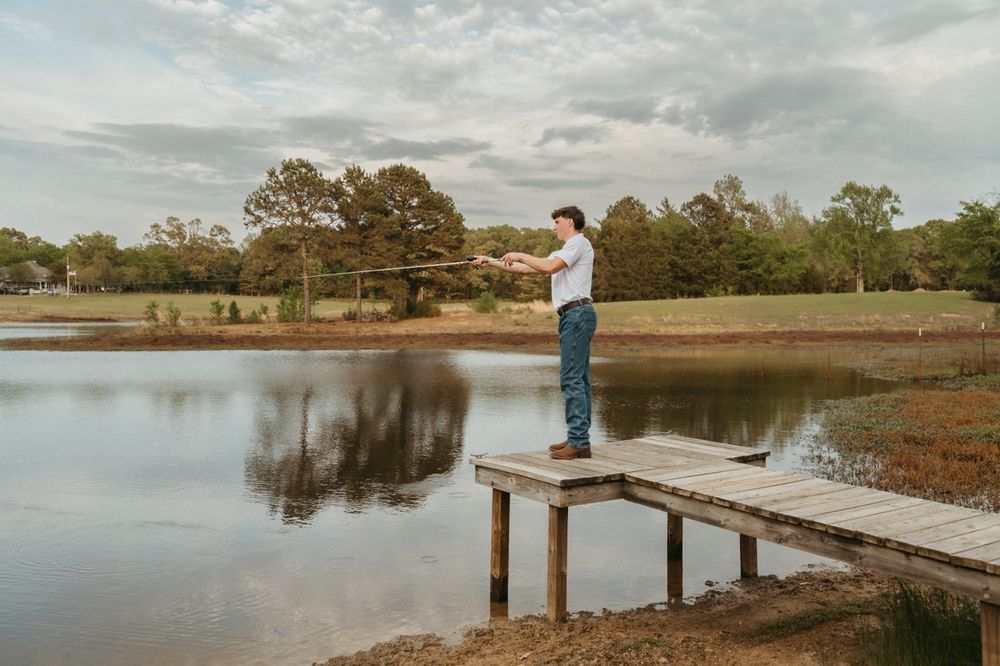 Man fishing from a wooden dock on a calm reflective lake surrounded by green trees under a cloudy sky.