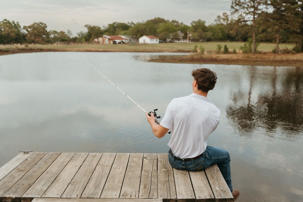 Young man in white t-shirt fishing from a wooden dock on a calm lake surrounded by trees.