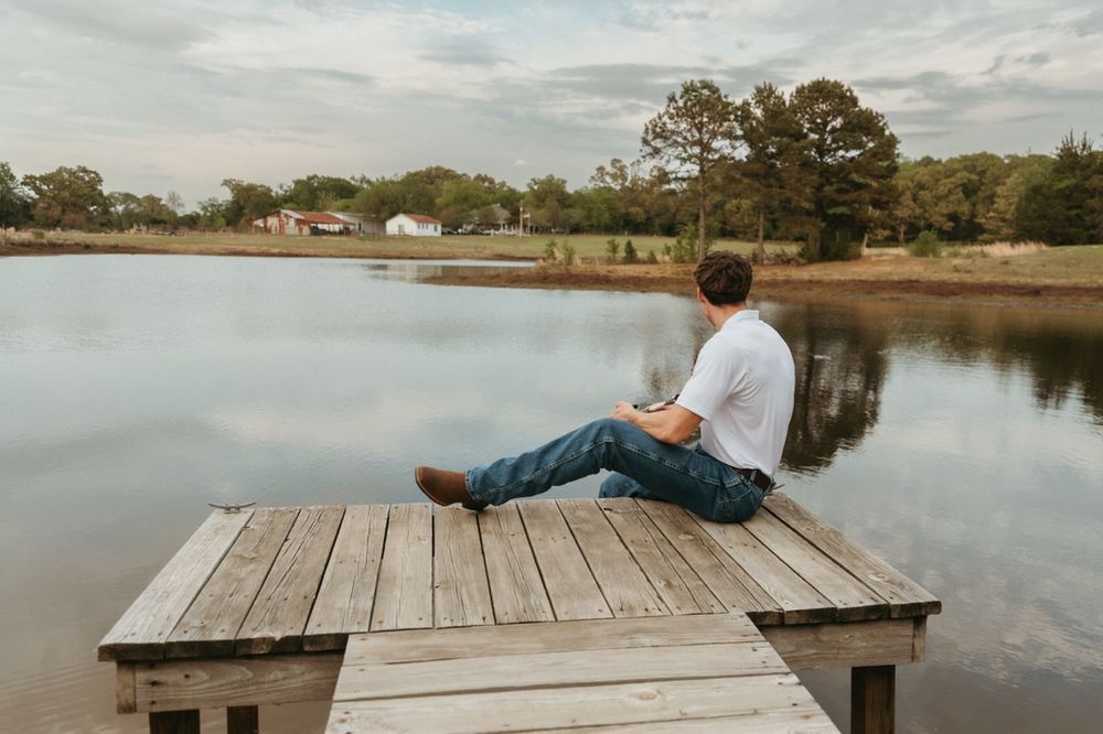 Young man sitting on wooden dock overlooking calm lake surrounded by trees on cloudy day.