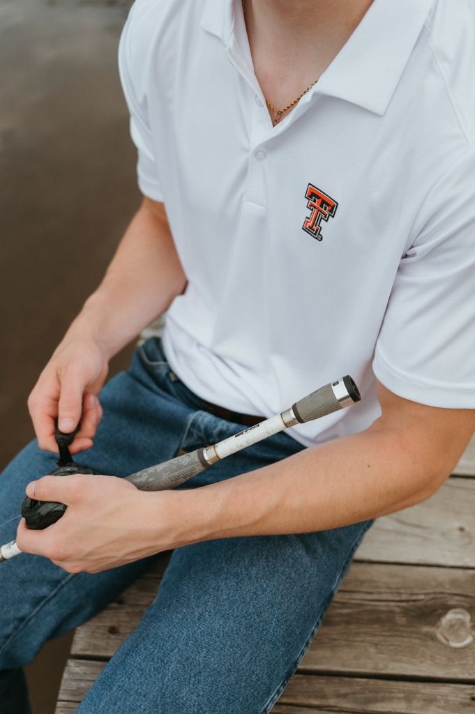 Man wearing white Texas Tech polo shirt sitting on wooden dock holding a prosthetic arm component.