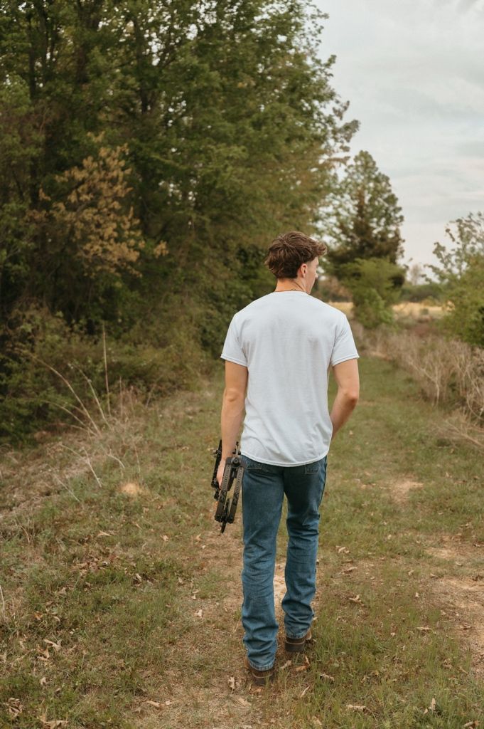 Young man in white t-shirt and jeans walks down a rural path carrying a guitar case surrounded by trees.