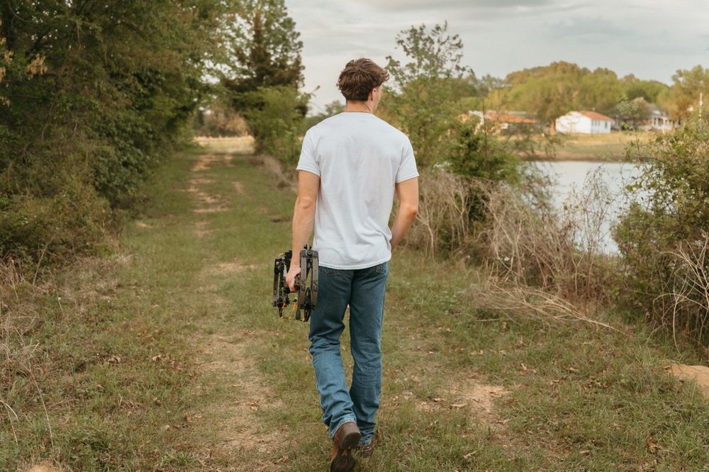 Young man walking a grassy path near a lake carrying a bow on a cloudy day.