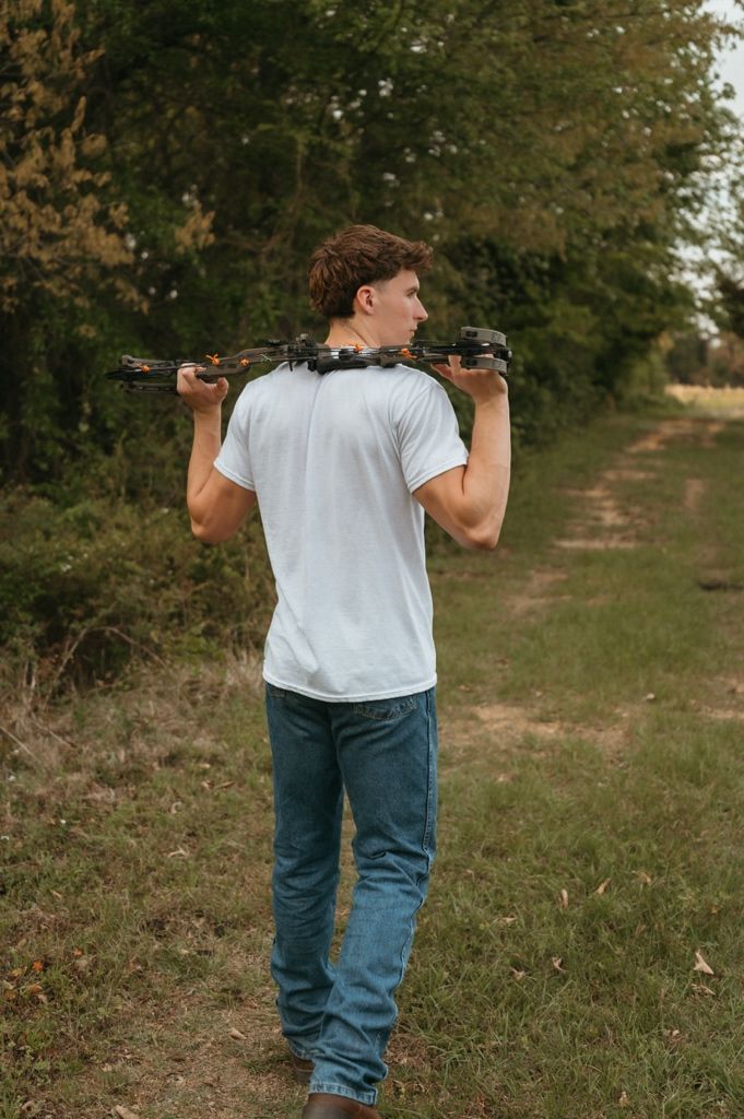 Young man carrying a drone on his shoulders while walking along a rural path surrounded by green trees.