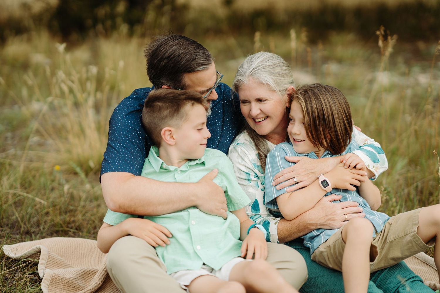 Family of four sitting outdoors in a field, sharing a warm embrace and joyful smiles together during a photography session
