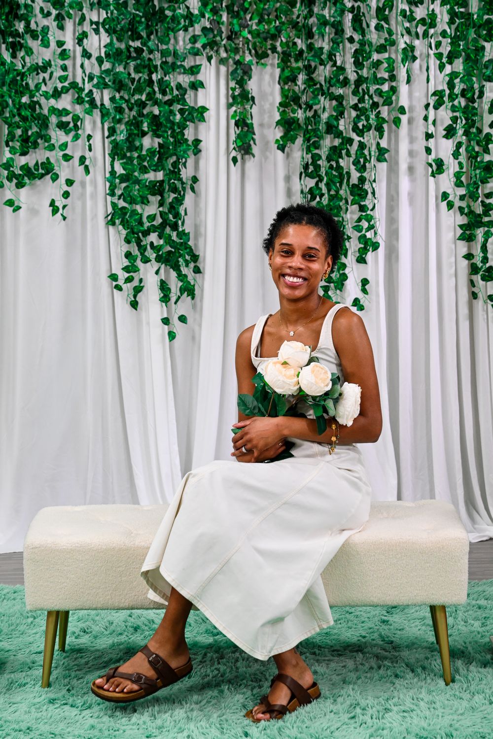 Smiling woman in white dress holding white roses, seated on bench with green ivy backdrop.