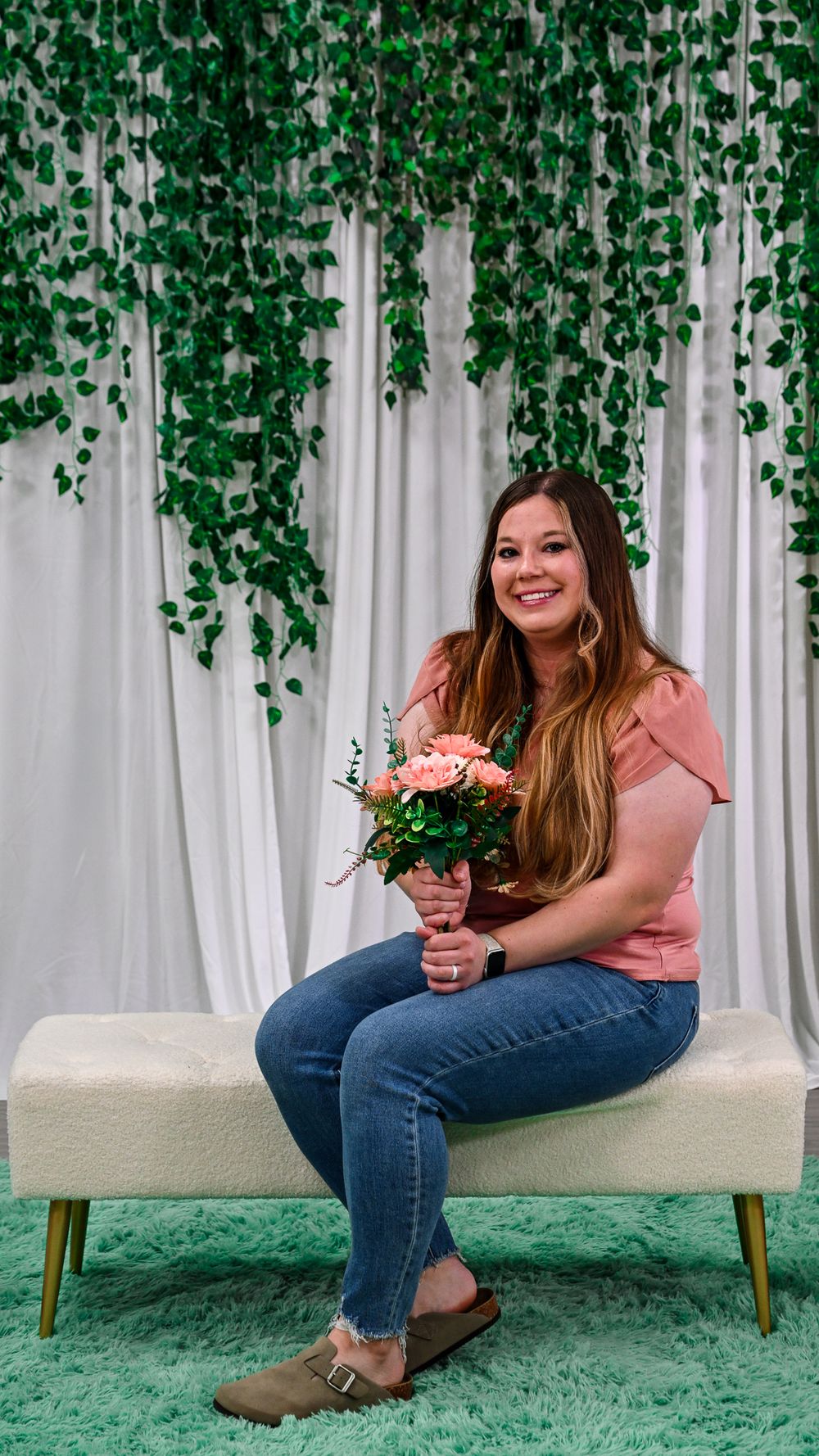 Woman in pink top and jeans sits on white bench holding pink roses with green ivy backdrop.