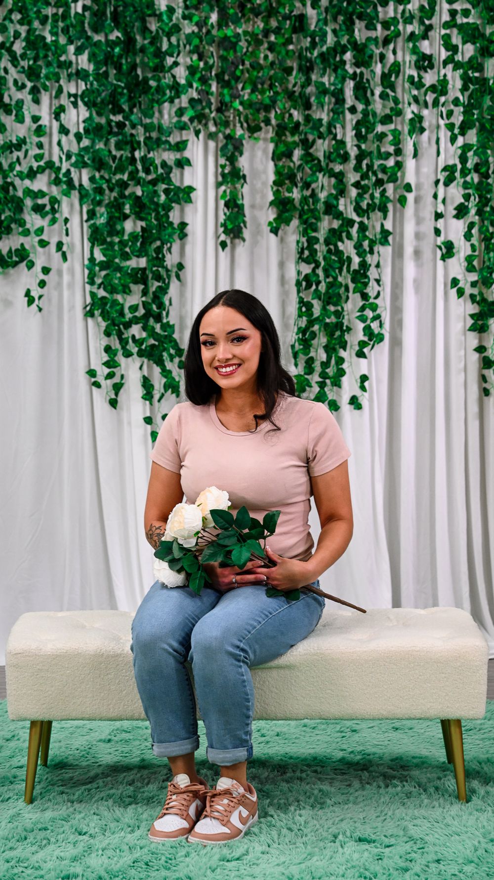 Woman in beige top and jeans sits on white bench holding white roses with ivy curtain backdrop.