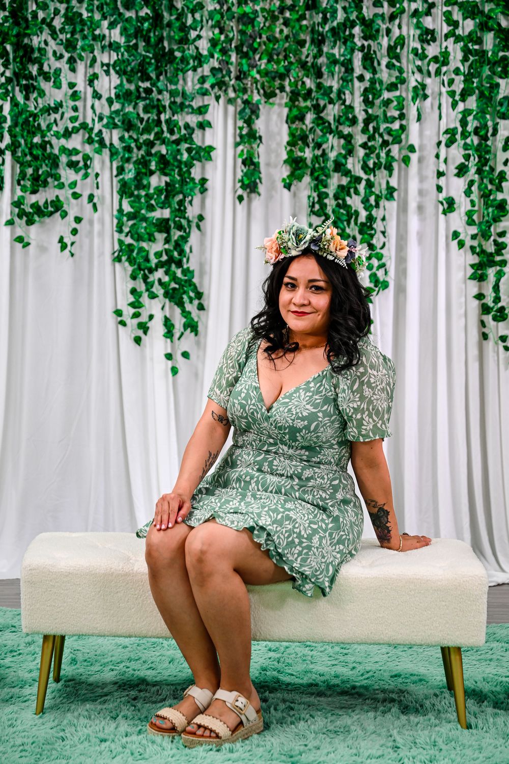 Woman in green floral dress with flower crown sits on white bench before hanging vine backdrop.