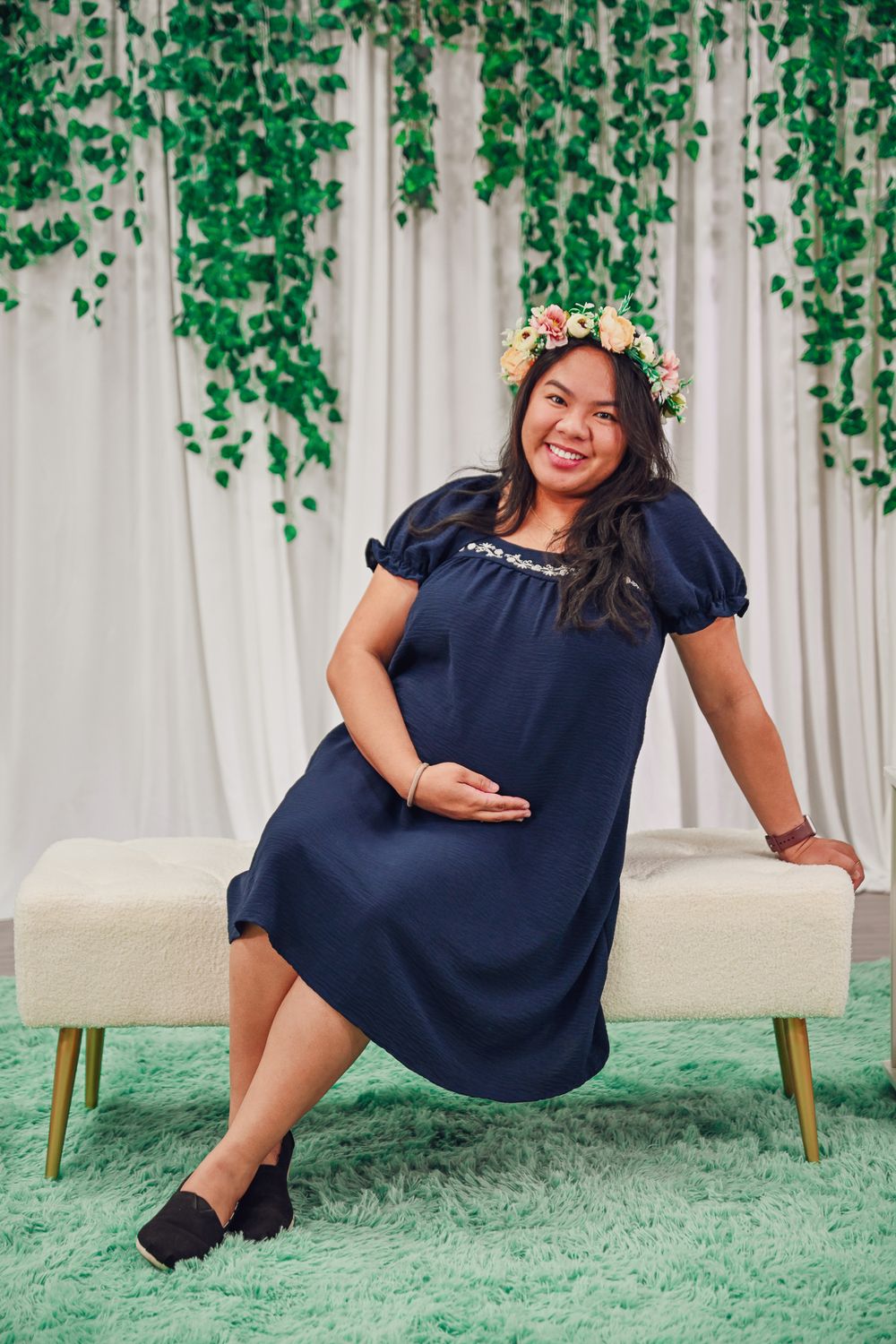 Smiling pregnant woman in navy blue dress with floral crown poses on bench with greenery backdrop.
