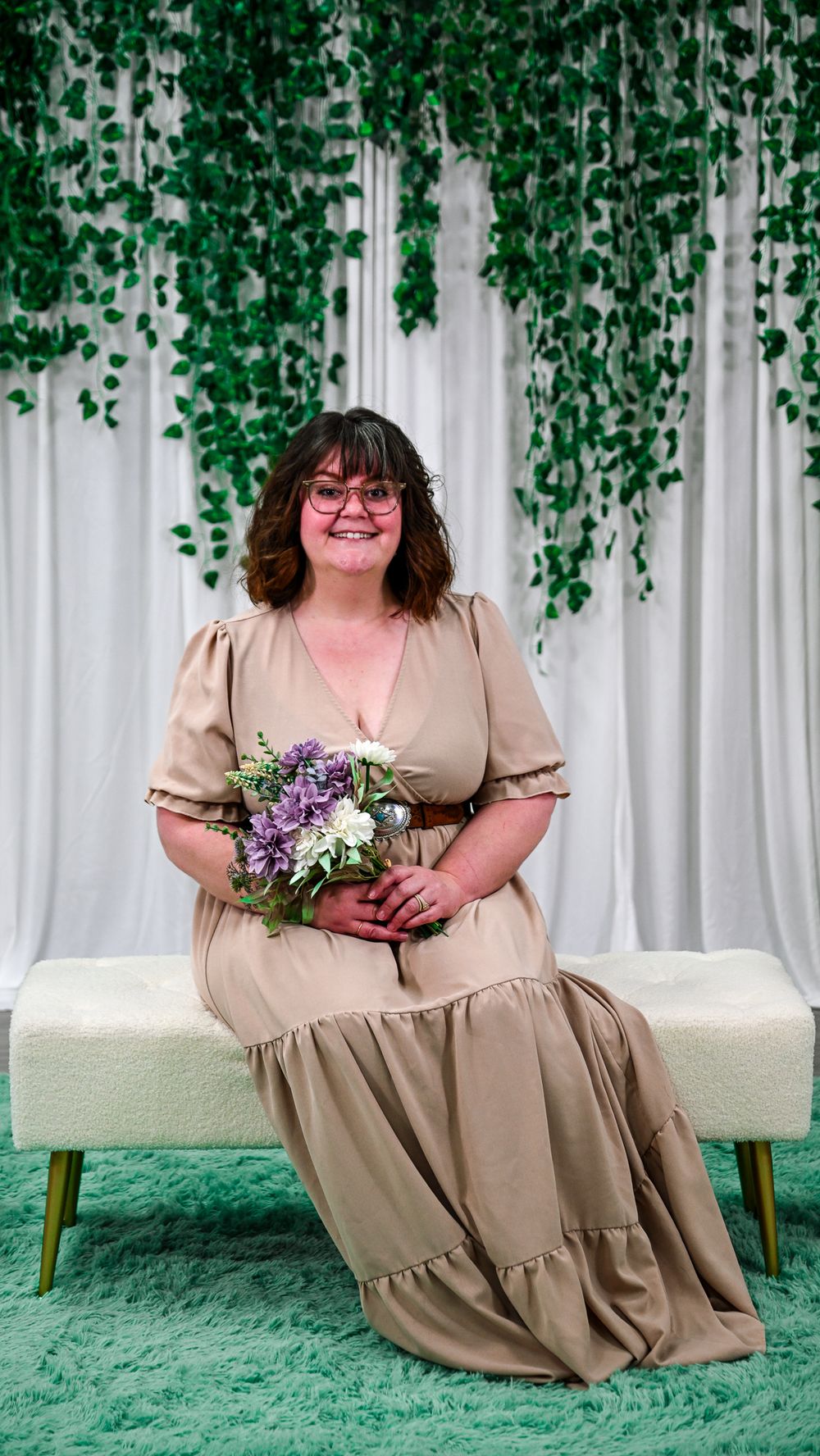 Woman in tan maxi dress holding purple bouquet sits on bench before white curtain with hanging greenery.