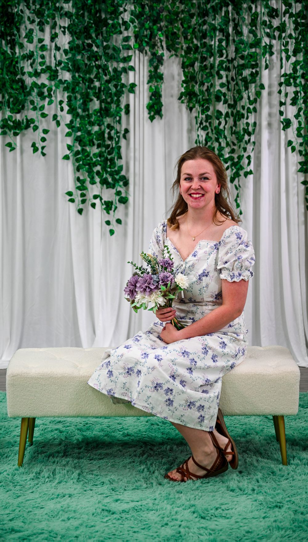 Woman in floral dress holding purple bouquet sits on bench with green ivy backdrop.