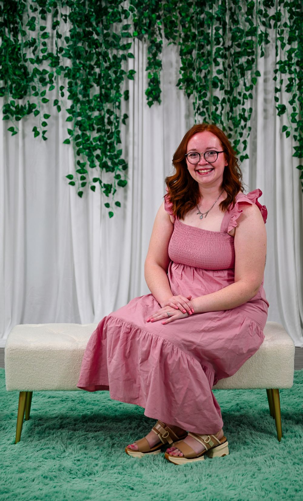 Redhead woman in pink maxi dress with glasses sits smiling on white bench with ivy backdrop.